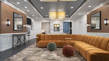 Un moderno vestíbulo de hotel con un amplio sofá modular, un taburete redondo, una alfombra estampada y un área de comedor con mesa y sillas.