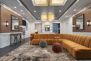 Un moderno vestíbulo de hotel con un amplio sofá modular, un taburete redondo, una alfombra estampada y un área de comedor con mesa y sillas.