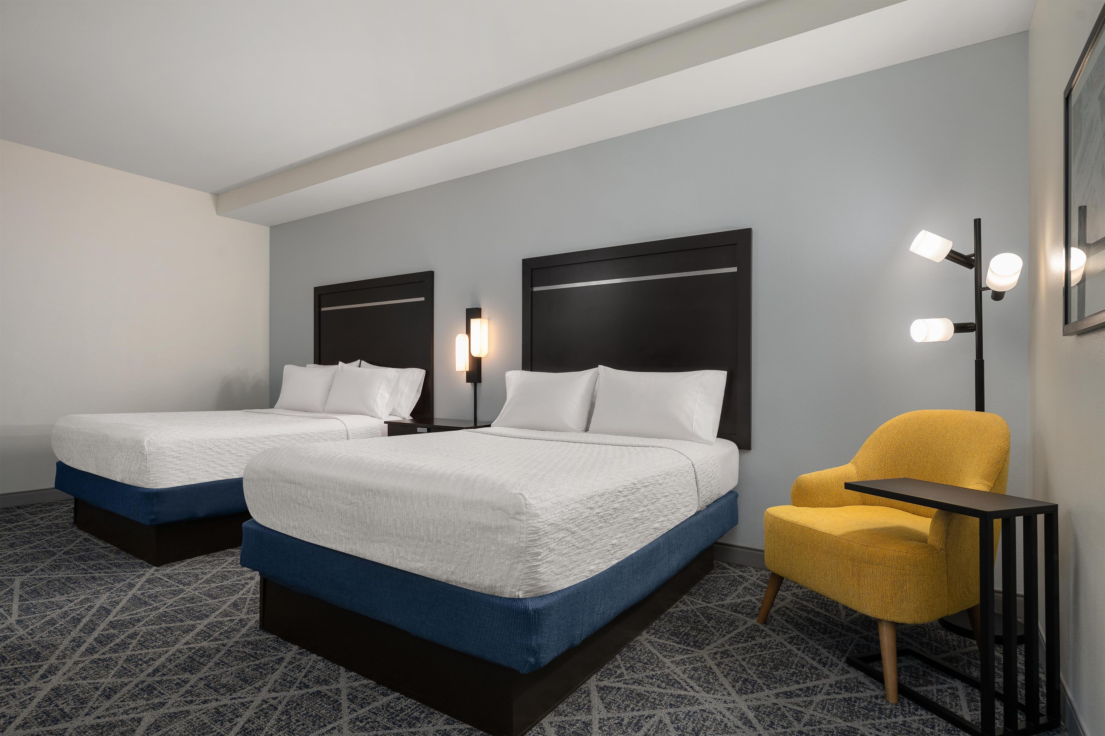 A hotel room with two beds, a yellow armchair, a small table, and a wall-mounted light fixture.