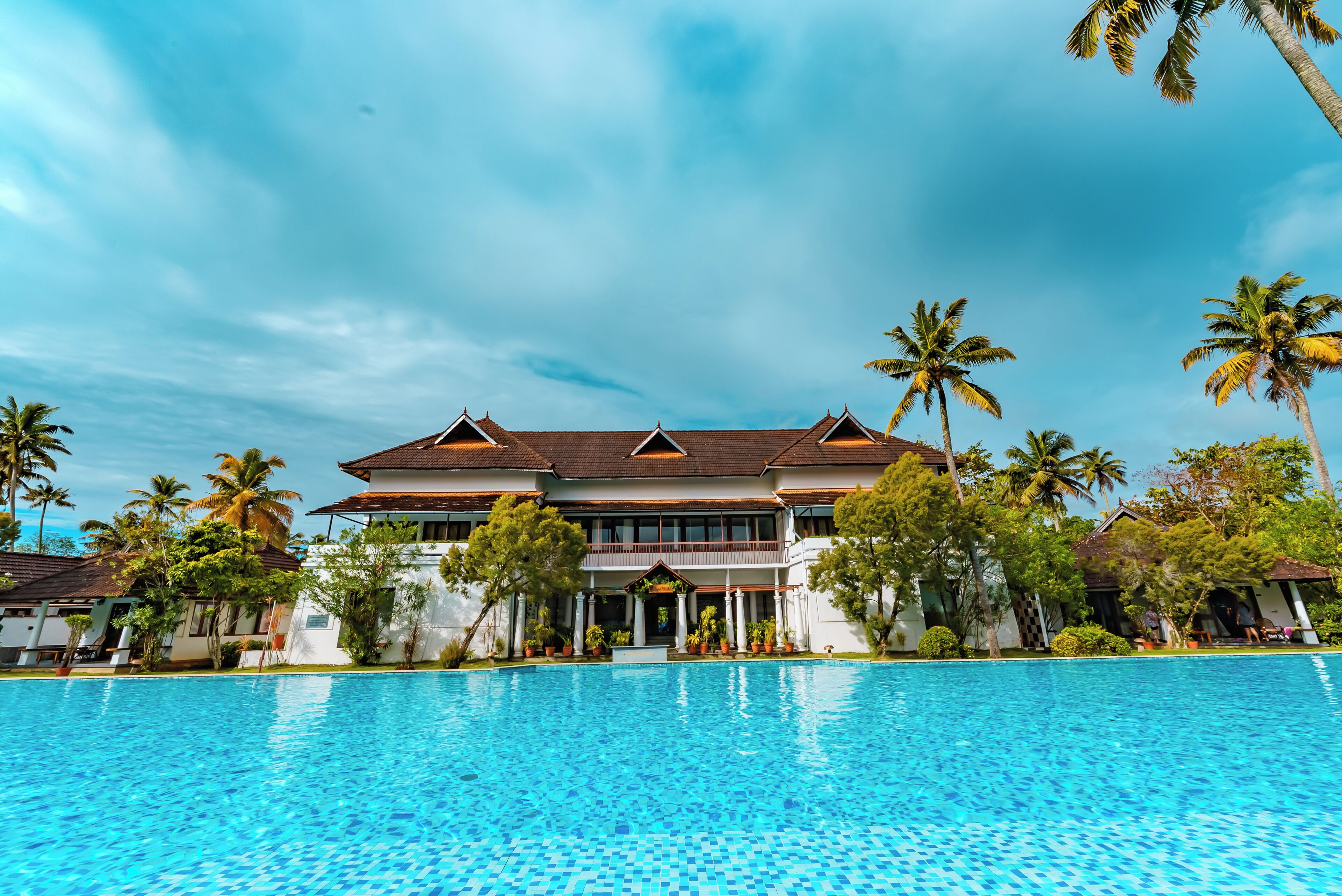 A large hotel with a swimming pool, palm trees, and a clear blue sky.