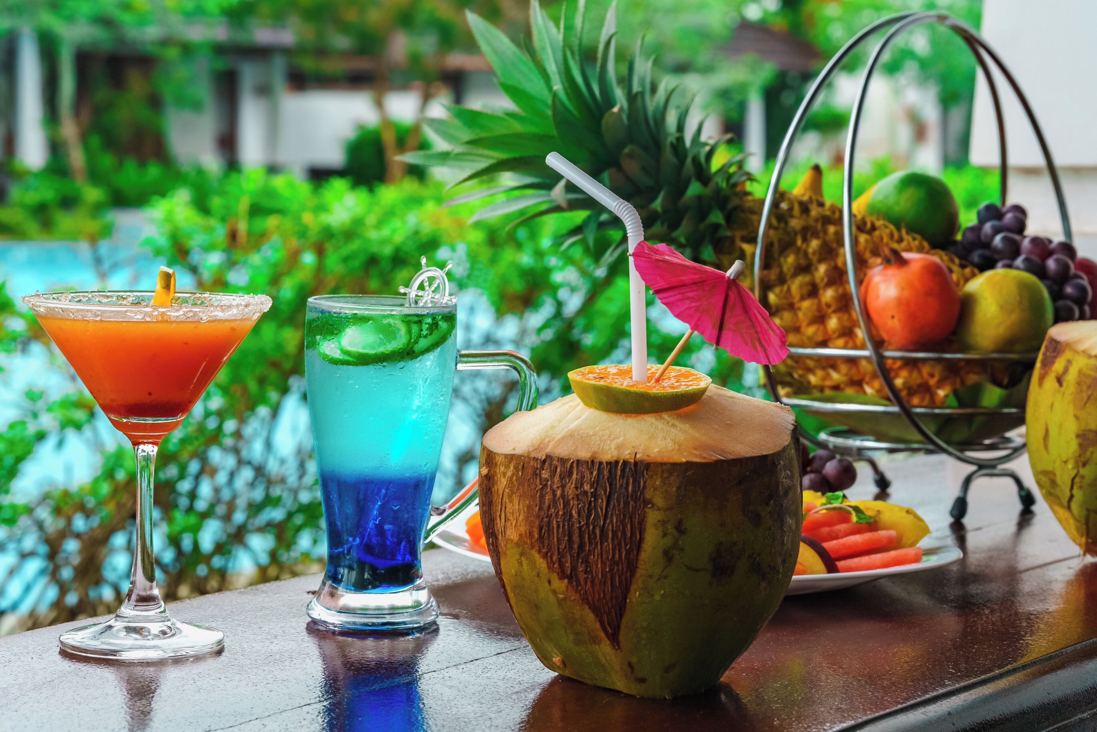 Disposición de bebidas tropicales con una copa de martini, un cóctel azul y una bebida de coco, junto con una cesta de frutas y una piña.