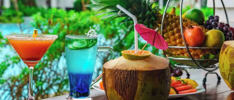 Disposición de bebidas tropicales con una copa de martini, un cóctel azul y una bebida de coco, junto con una cesta de frutas y una piña.