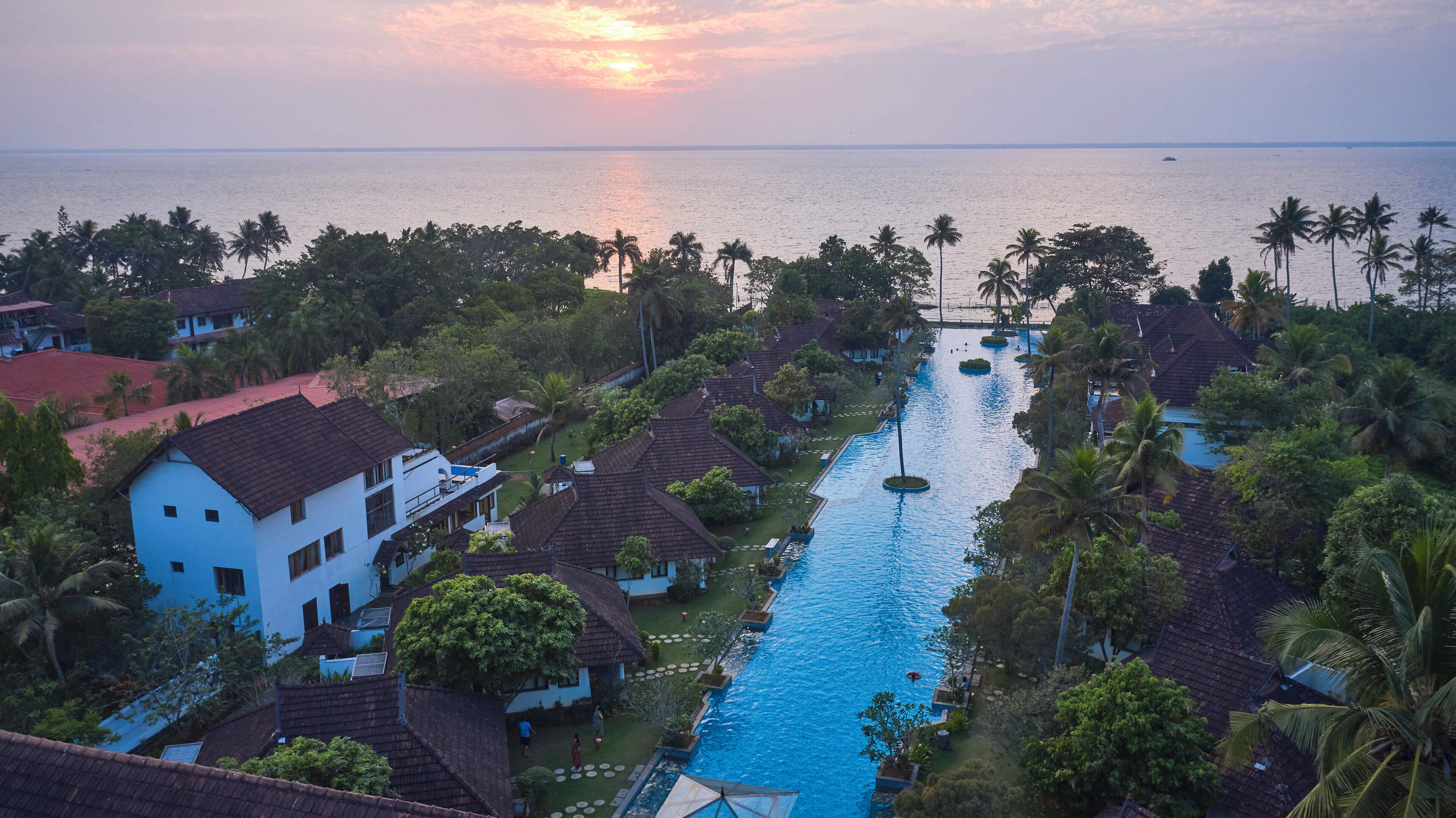 A resort with a swimming pool, palm trees, and a view of the sea at sunset.