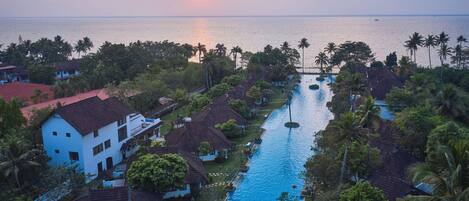 A resort with a swimming pool, palm trees, and a view of the sea at sunset.