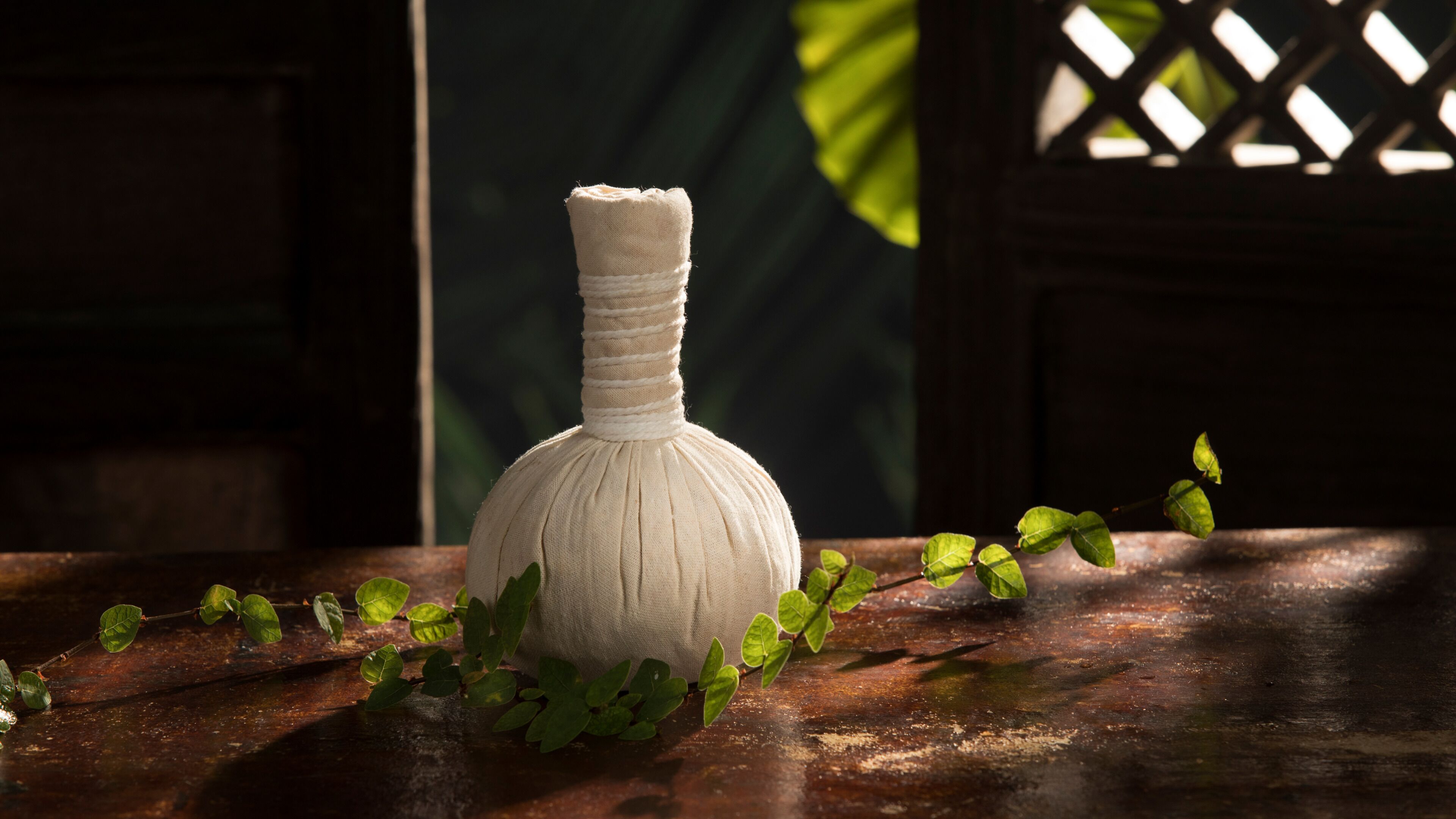 Un jarrón blanco con superficie texturizada sobre una mesa de madera, rodeado de hojas verdes.