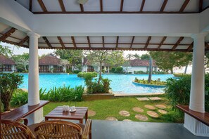 A covered outdoor area with a pool, wooden furniture, and a view of villas in the background. - Rhythm Kumarakom (Kottayam)