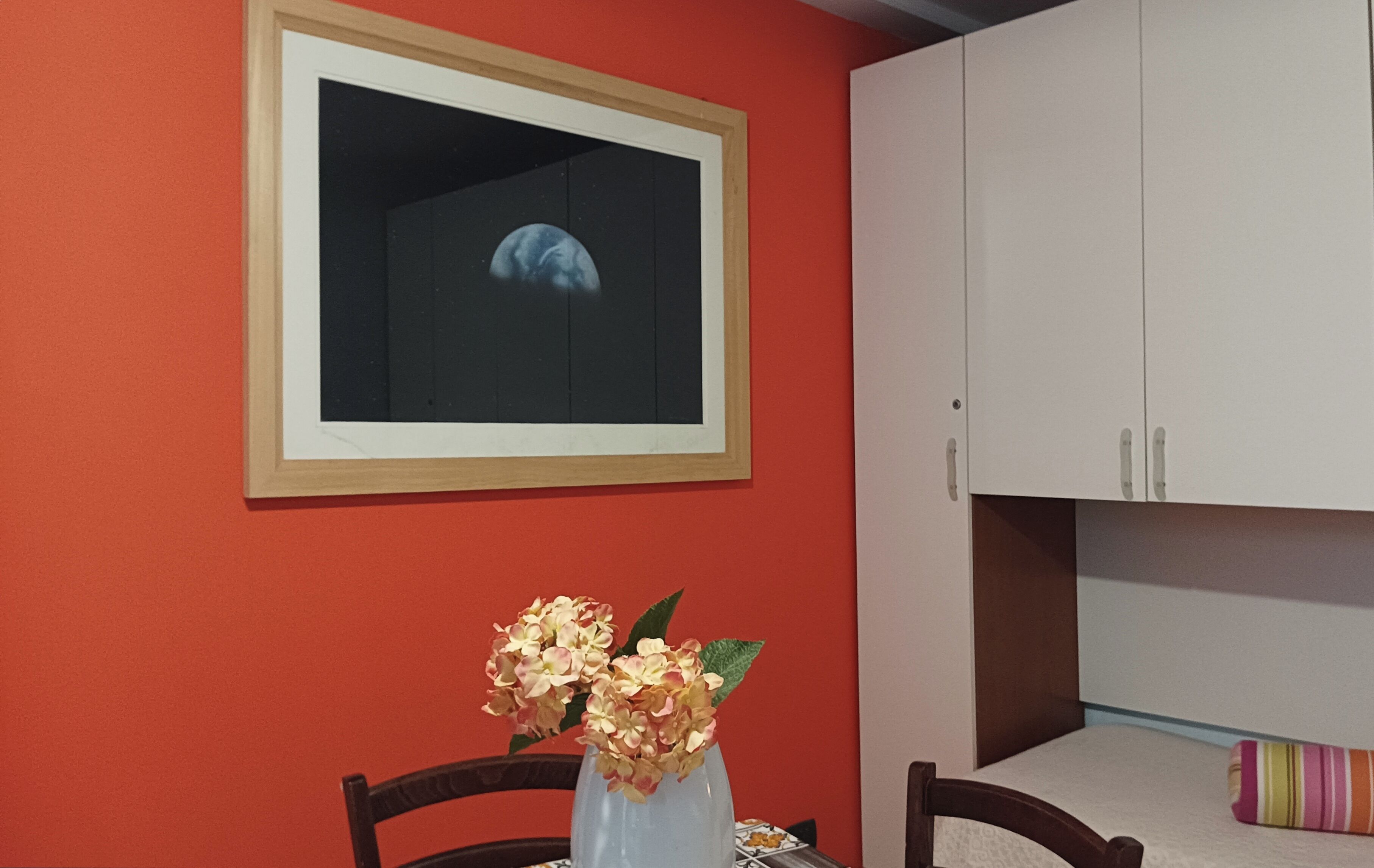 Una habitación con una pared roja, un cuadro enmarcado, un armario blanco y una mesa con un florero.