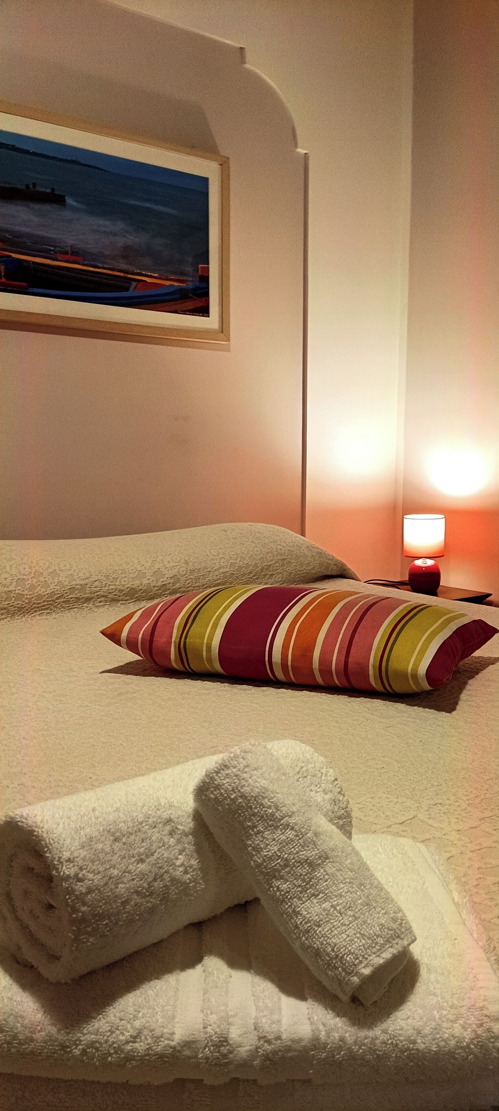 A neatly made bed with a striped pillow, a rolled white towel, and a painting of a seascape on the wall.