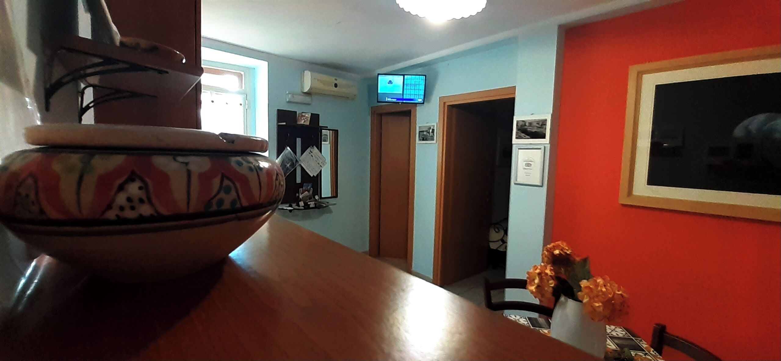 Un coin repas avec une table en bois, un bol décoratif et un vase rempli de fleurs. Un téléviseur est fixé au mur, et une porte donne accès à une autre pièce.