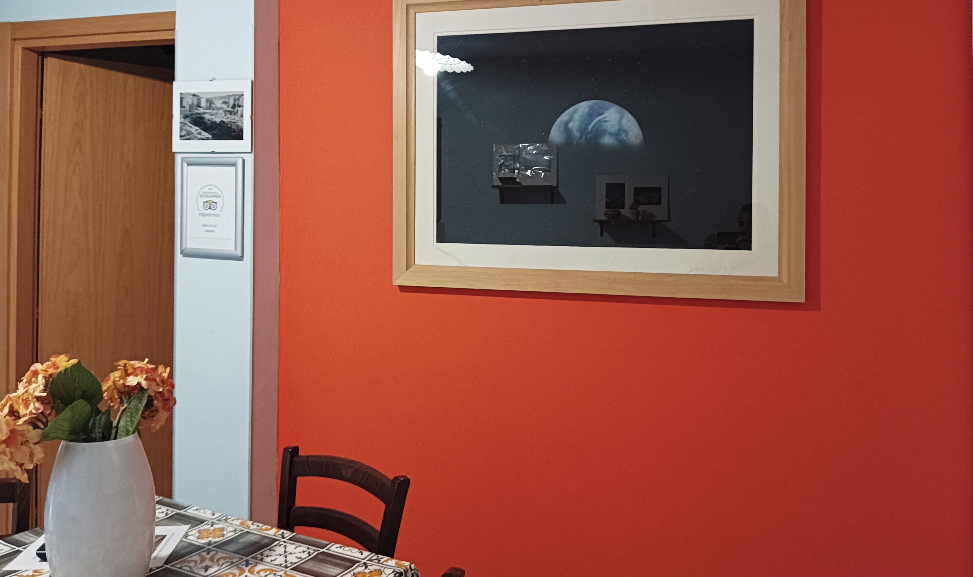 A dining area with a table set for two, a vase with flowers, and a framed picture on the wall.