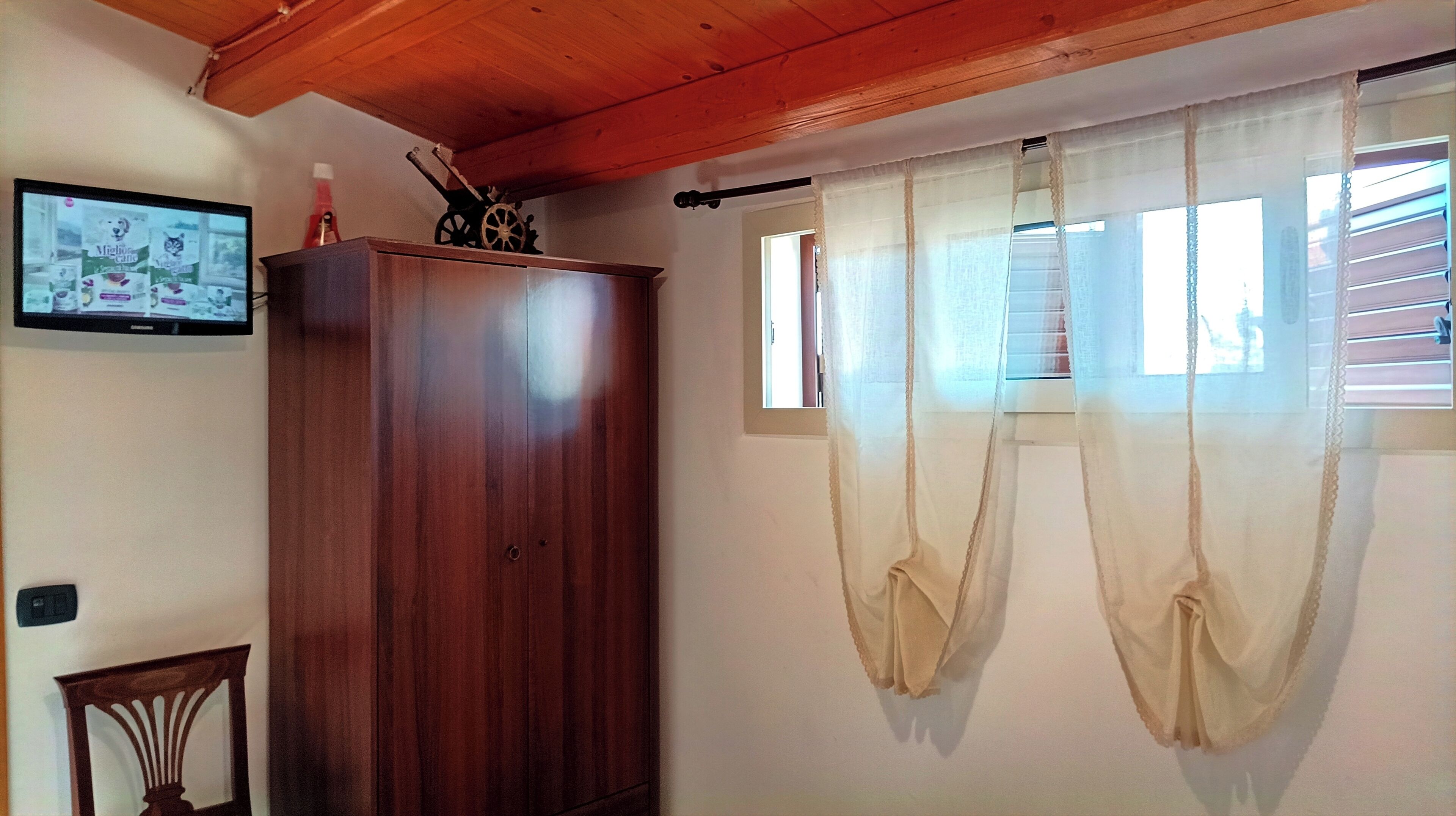 Una habitación con un armario de madera, un televisor montado en la pared y cortinas traslúcidas sobre una ventana.