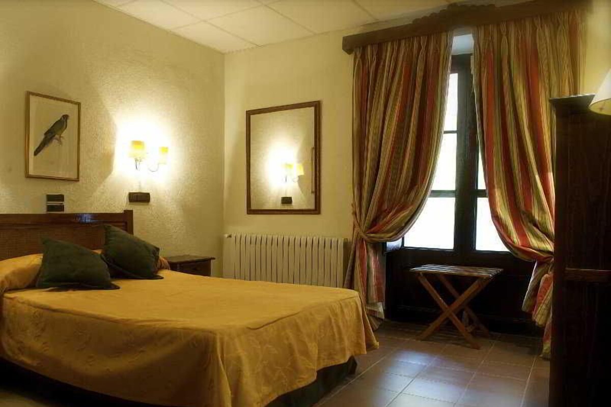 A hotel room with a bed, a small table, a radiator, and curtains.