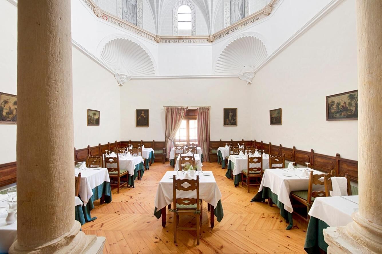 A dining hall with wooden tables and chairs, white tablecloths, and a high arched ceiling.
