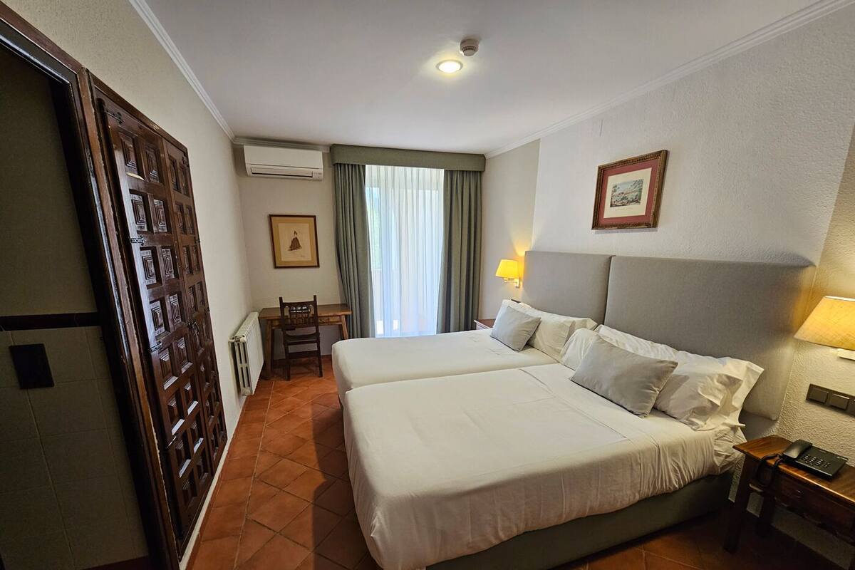 Habitación de hotel con dos camas, un escritorio, una silla, un equipo de aire acondicionado y una ventana con cortinas.