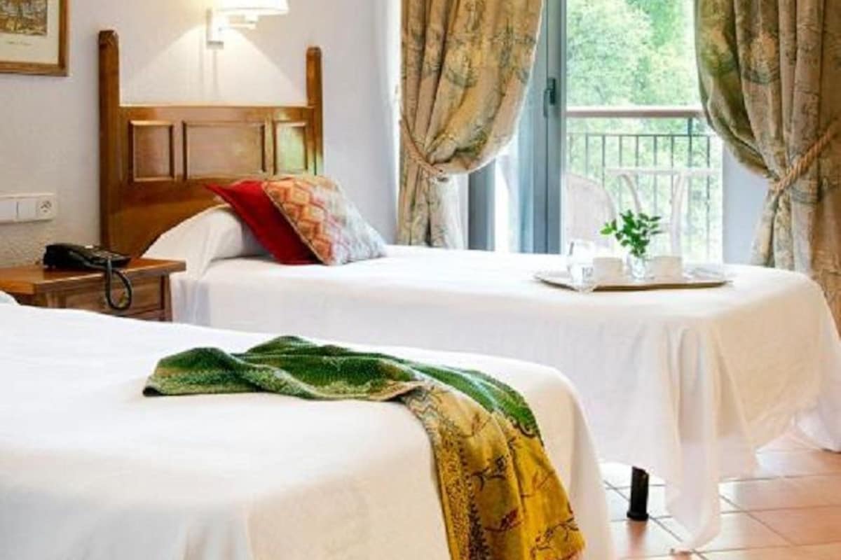 Een hotelkamer met twee bedden, een nachtkastje met een lamp, een raam met gordijnen en uitzicht op groen.
