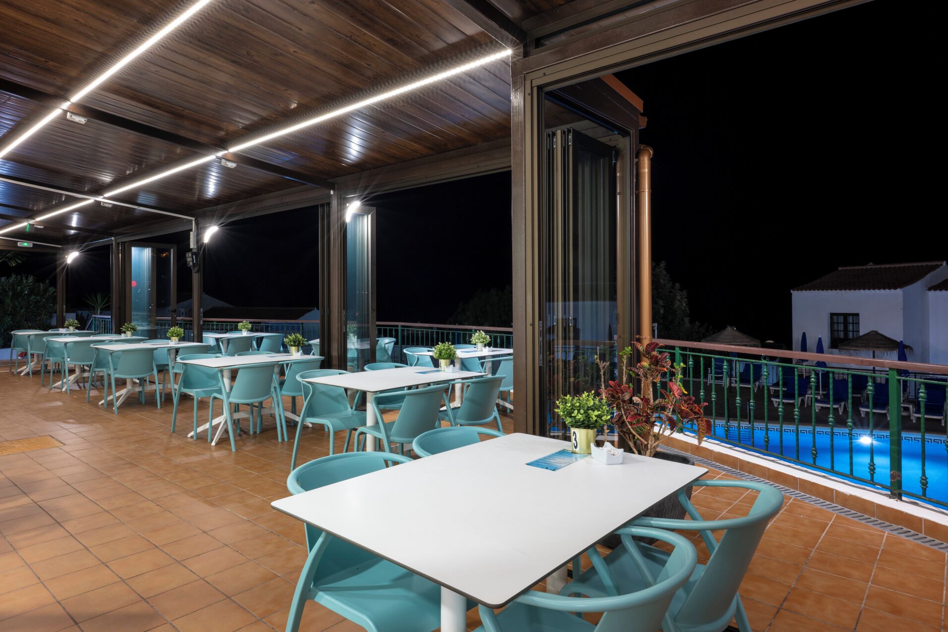 An outdoor dining area with tables and chairs, a pool in the background, and a covered section with a wooden ceiling.