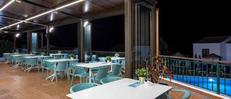 An outdoor dining area with tables and chairs, a pool in the background, and a covered section with a wooden ceiling.