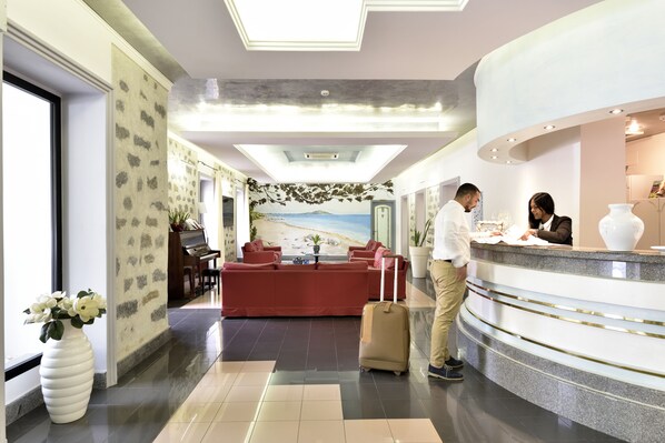 A hotel lobby with a reception desk, a red sofa, a piano, and a large window. - Hotel Orri (Tortoli)