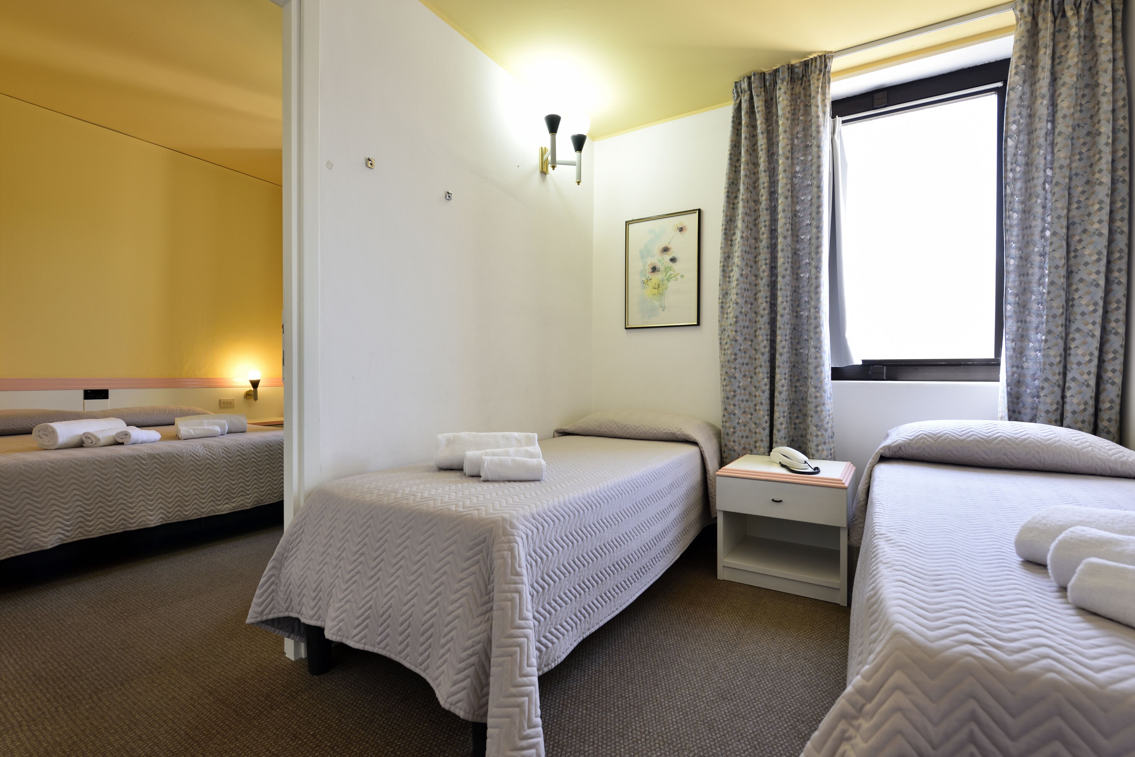 A hotel room with two beds, a bedside table with a lamp, a framed picture on the wall, and a window with curtains.