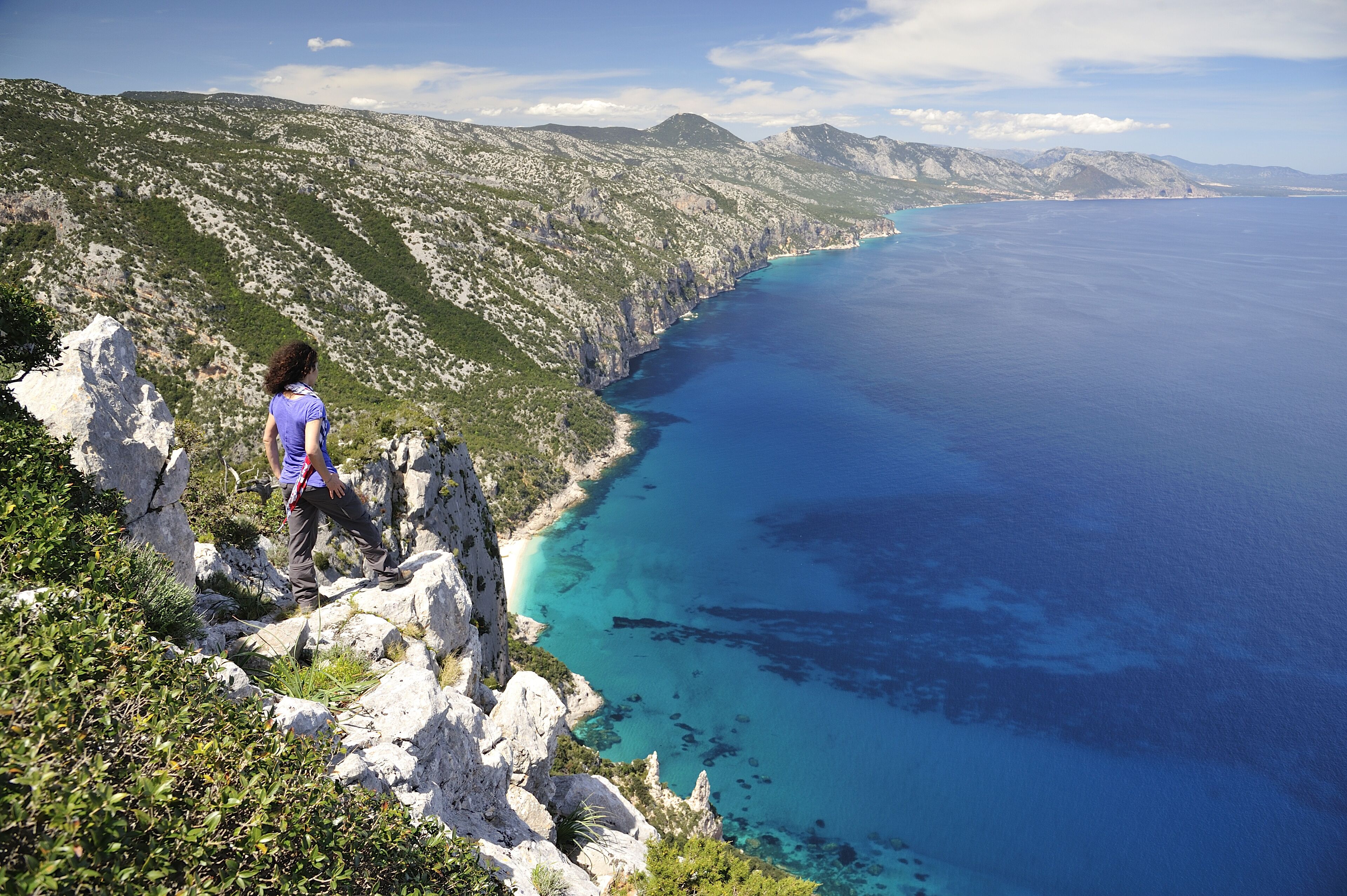 Une personne qui fait de la randonnée sur un sentier côtier rocheux, avec vue sur la mer et les montagnes.