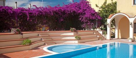 A swimming pool with steps leading to a covered area, surrounded by purple flowering plants and a yellow building.