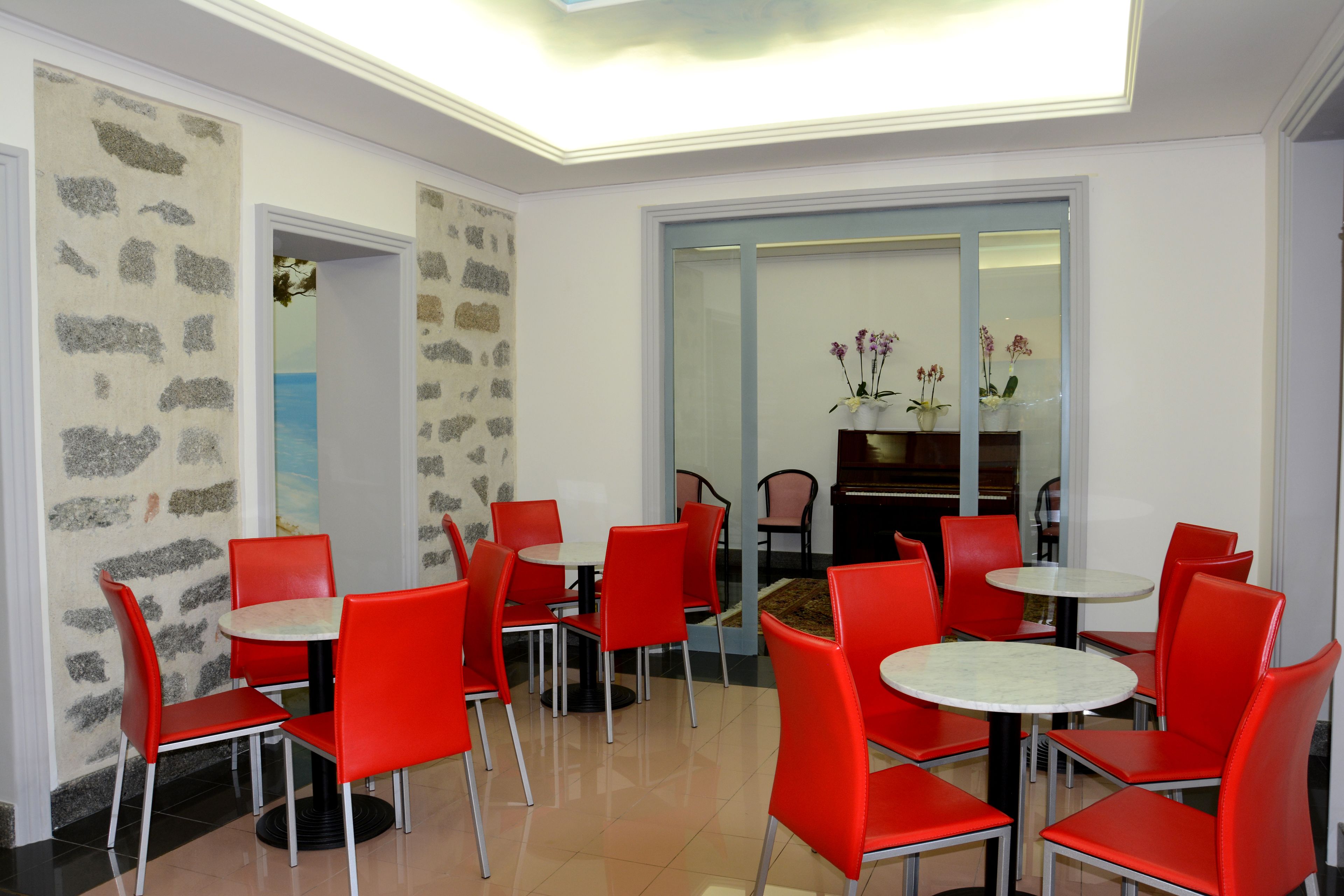 A dining area with red chairs and white tables, stone wall decor, and a view through a glass door.