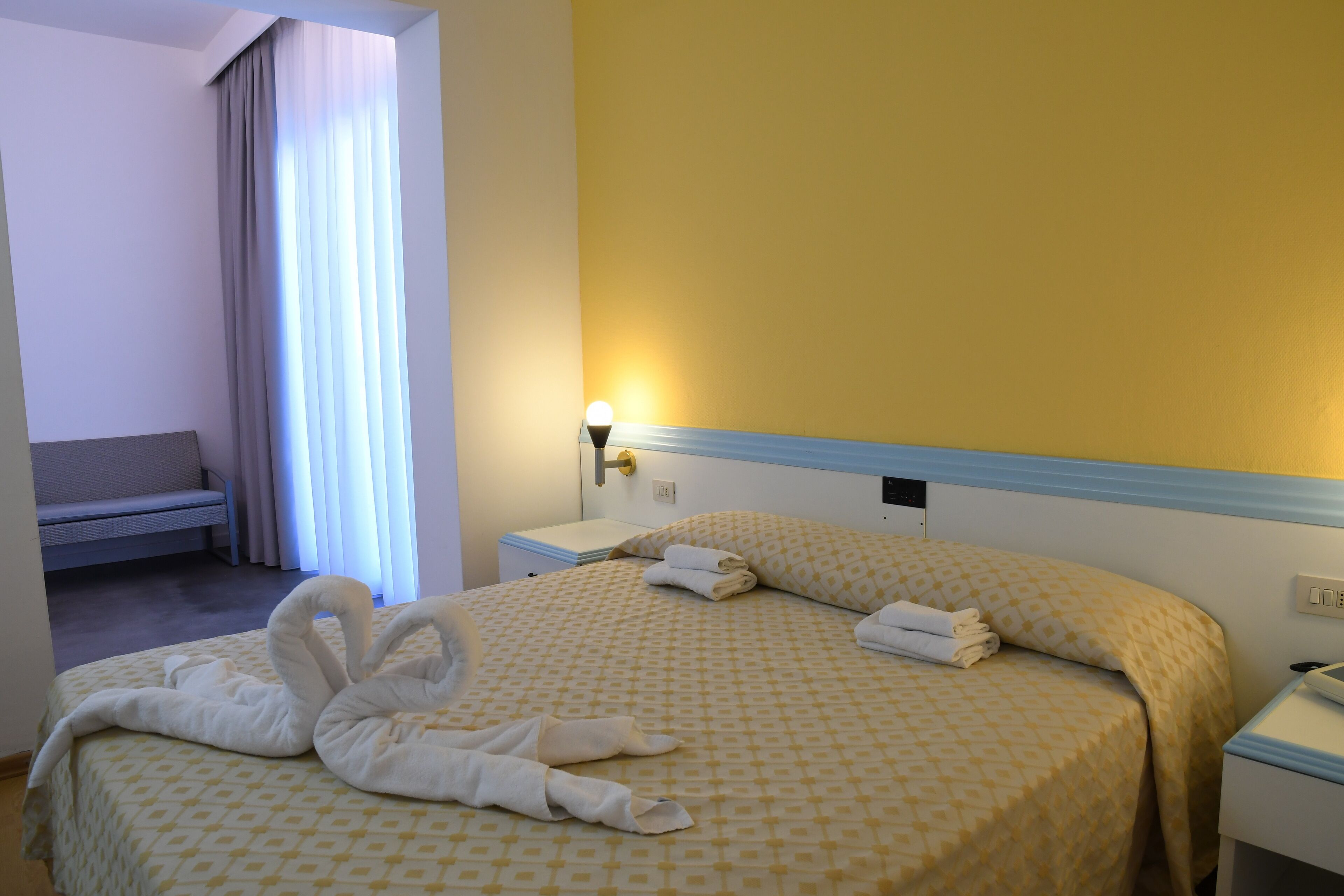 Quarto de hotel com uma cama, mesa de cabeceira, abajur e um banco.
