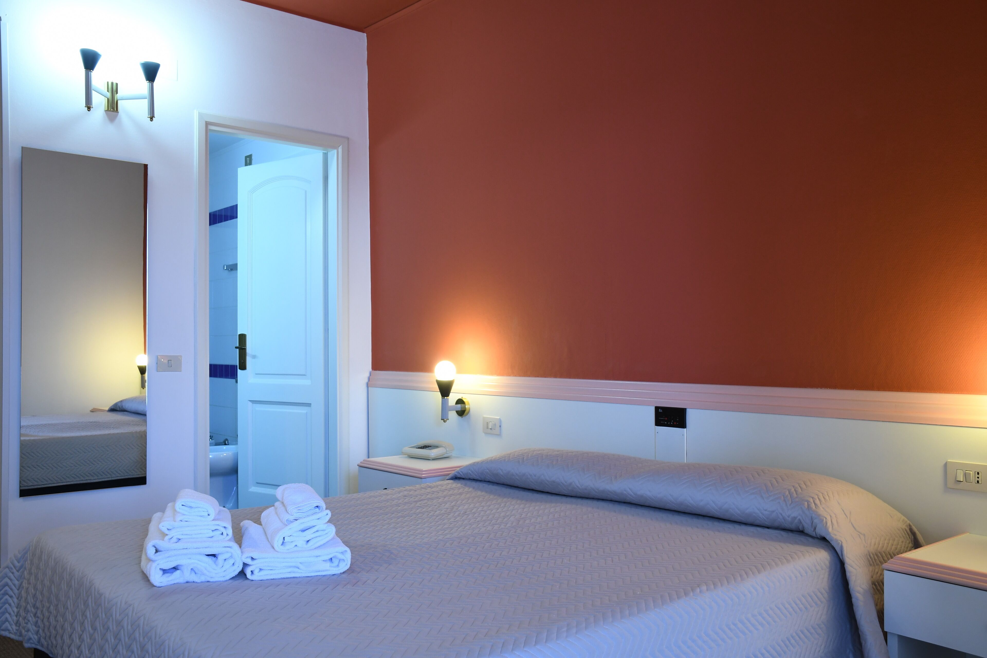 A hotel room with a bed, a nightstand, a mirror, and a bathroom visible through an open door.