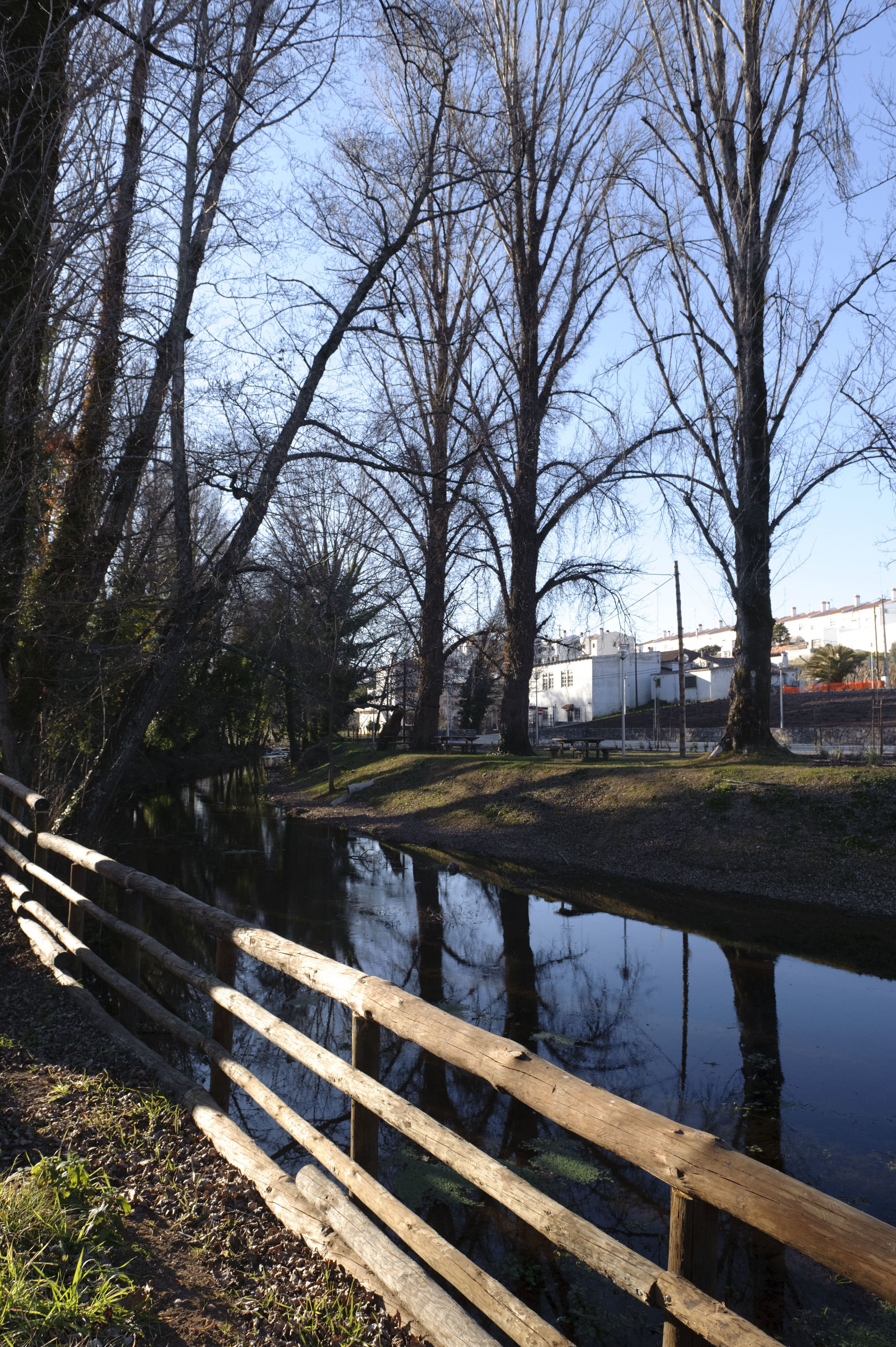 A wooden bridge over a calm river, surrounded by leafless trees and a building in the background.