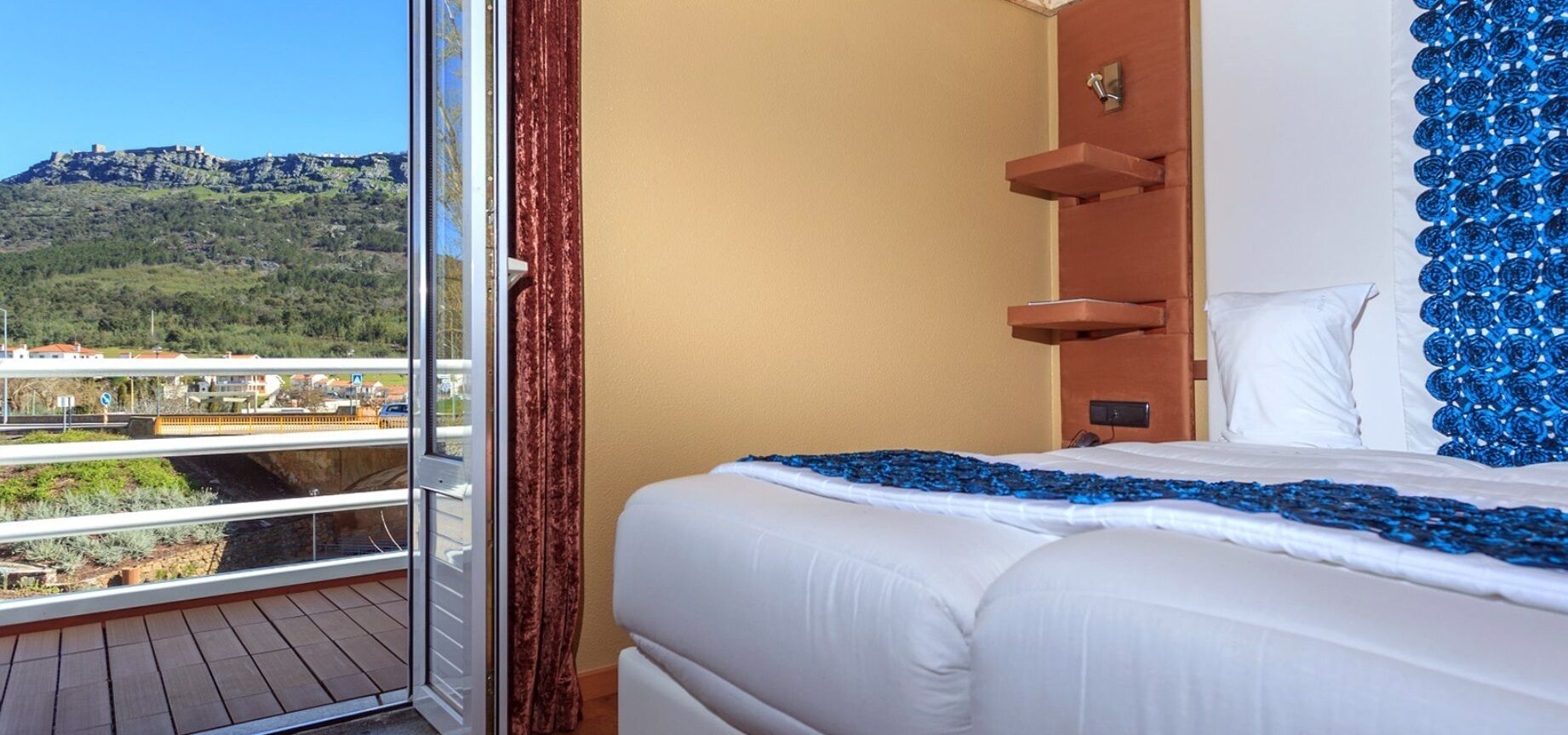 A hotel room with a bed, a window with a view of a mountain, and a balcony.