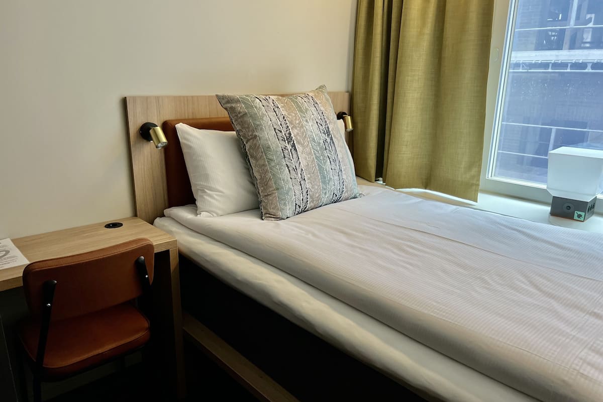Een enkel bed met houten hoofdeinde, een bureau met stoel, en een raam met gordijnen.