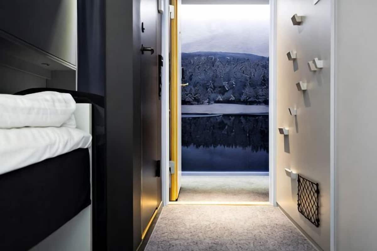 Een gang die leidt naar een slaapkamer met een bed, uitzicht op een meer en een muur met lichtschakelaars.