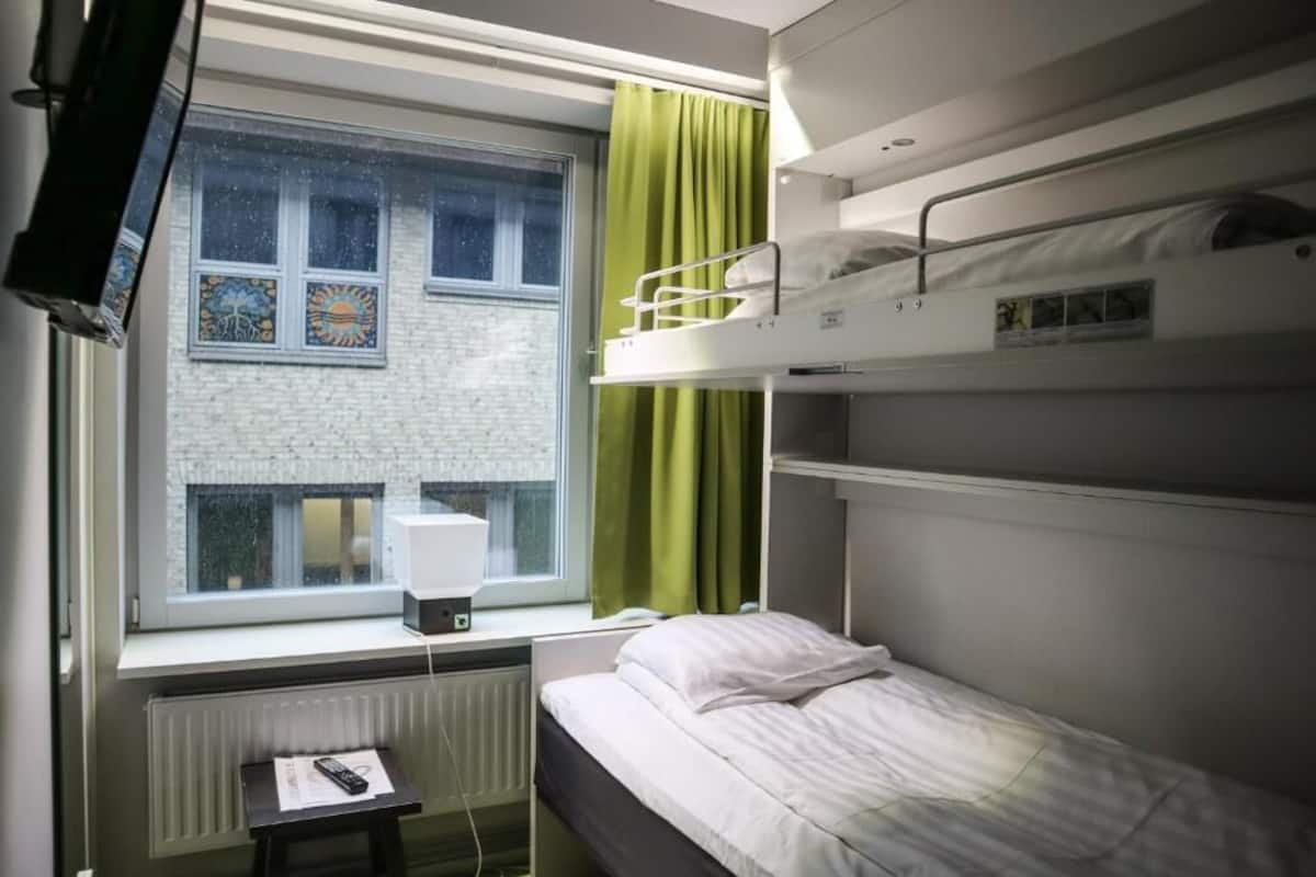 Een compacte hotelkamer met stapelbedden, een raam met gordijnen, een radiator en een klein tafeltje met een afstandsbediening.