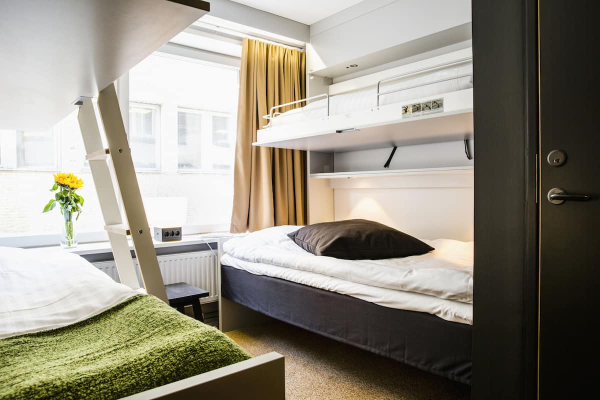 Een slaapzaal met stapelbedden, een raam met gordijnen, een radiator en een kleine tafel met een bloemenvaas.