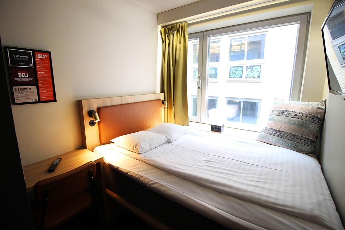 Habitación de hotel con una cama individual, una mesita de noche, un control remoto y una ventana con cortinas.