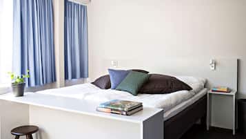 A modern bedroom with a bed, a bedside table with a book, a small stool, and a window with curtains.