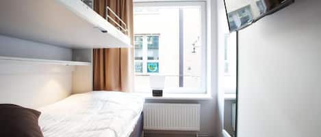 A hotel room with a bunk bed, a single bed, a window with curtains, a radiator, and a TV mounted on the wall.