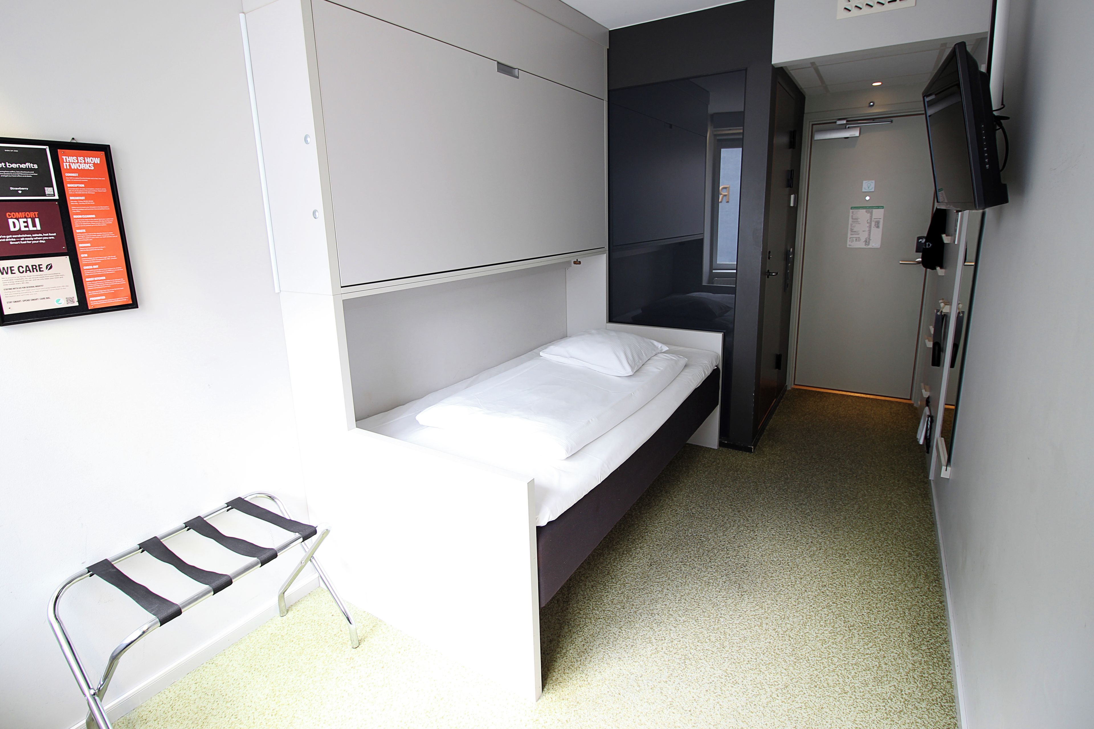 Une petite chambre d’hôtel étroite, avec un lit simple, une chaise et un bureau. Une porte donne accès à une autre pièce et un téléviseur est fixé au mur.