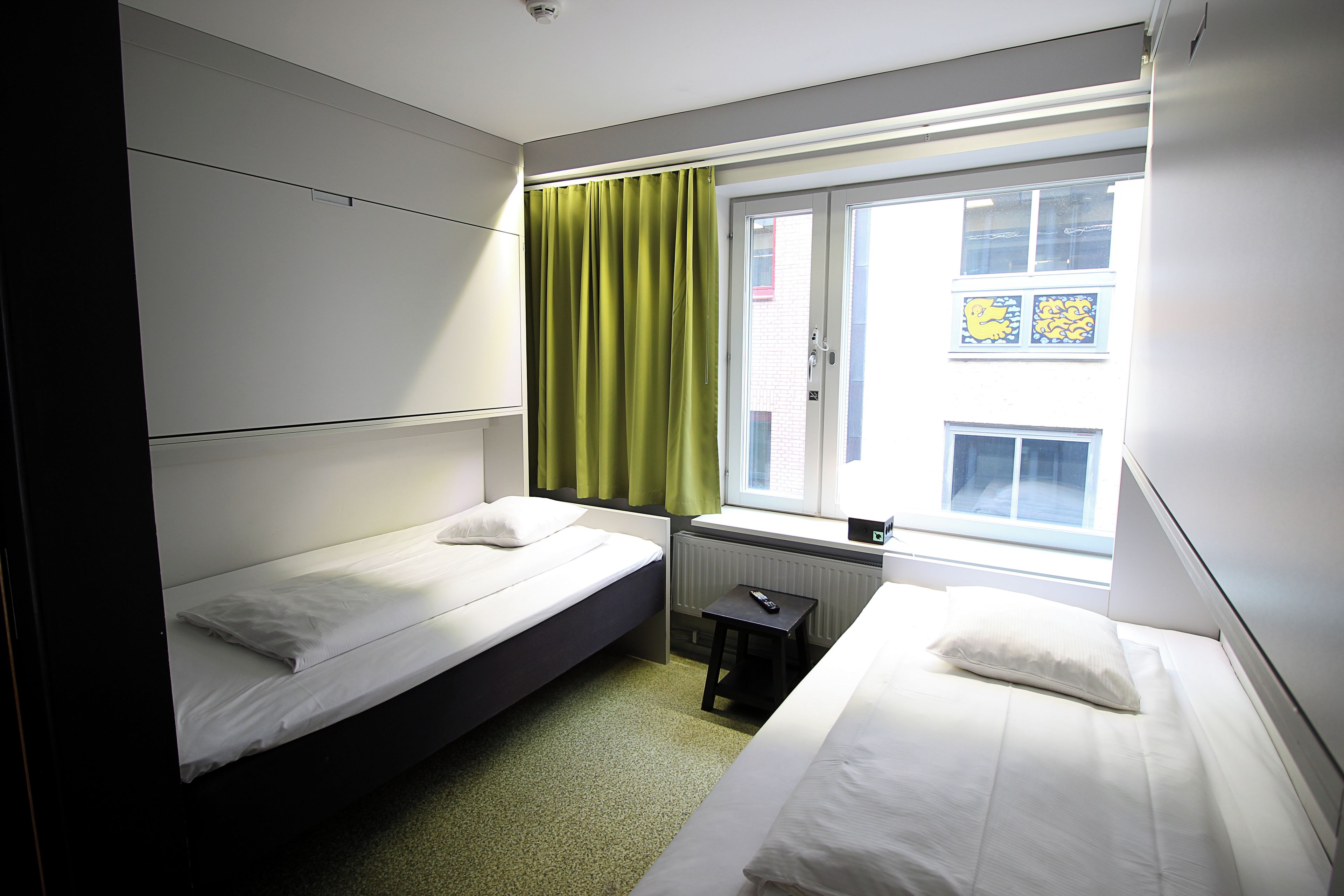 Ett hotellrum med två separata sängar, ett fönster med gröna gardiner, ett element och ett litet bord.