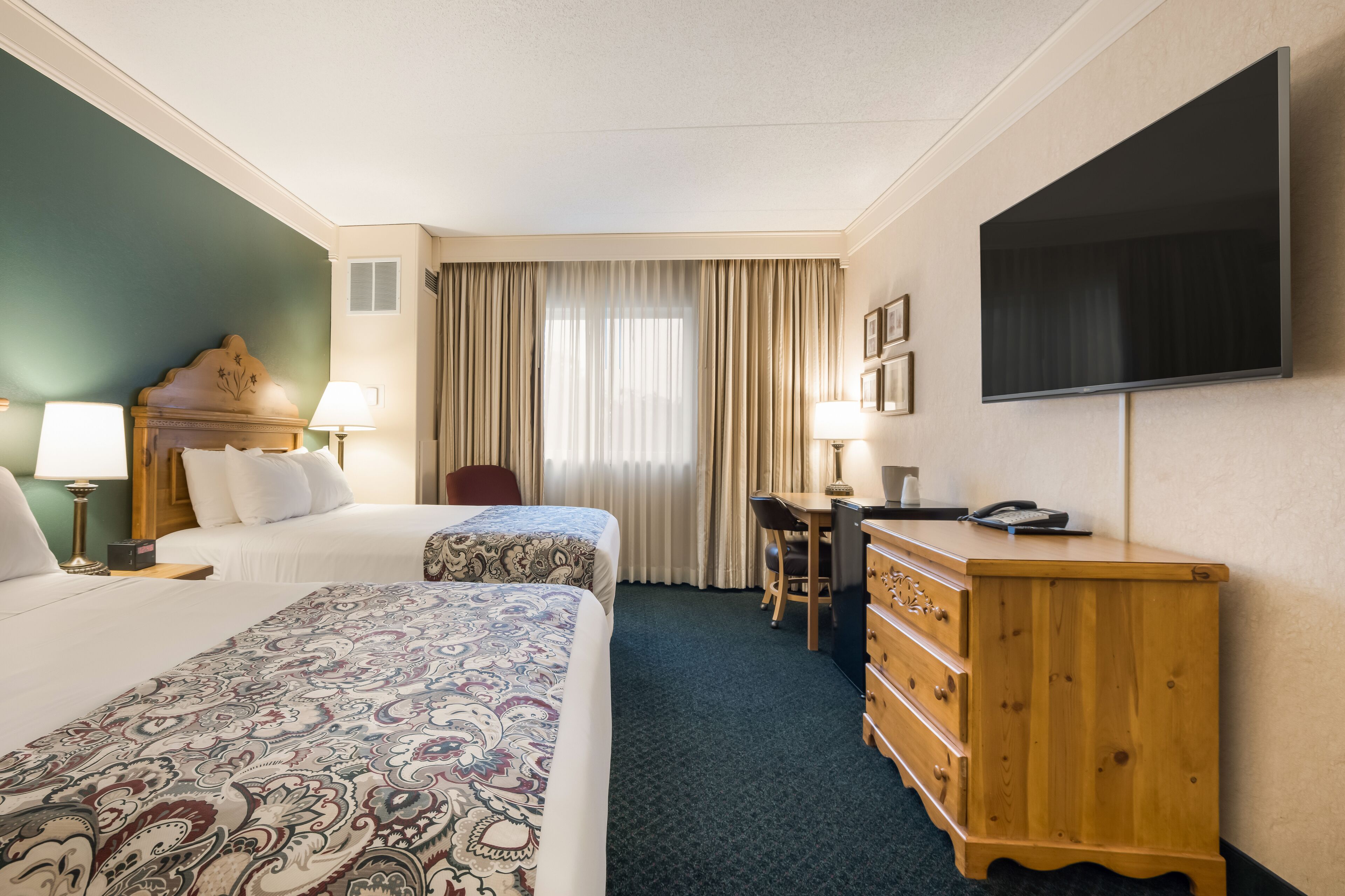 A hotel room with two beds, a desk, a dresser, a TV, and a window with curtains.