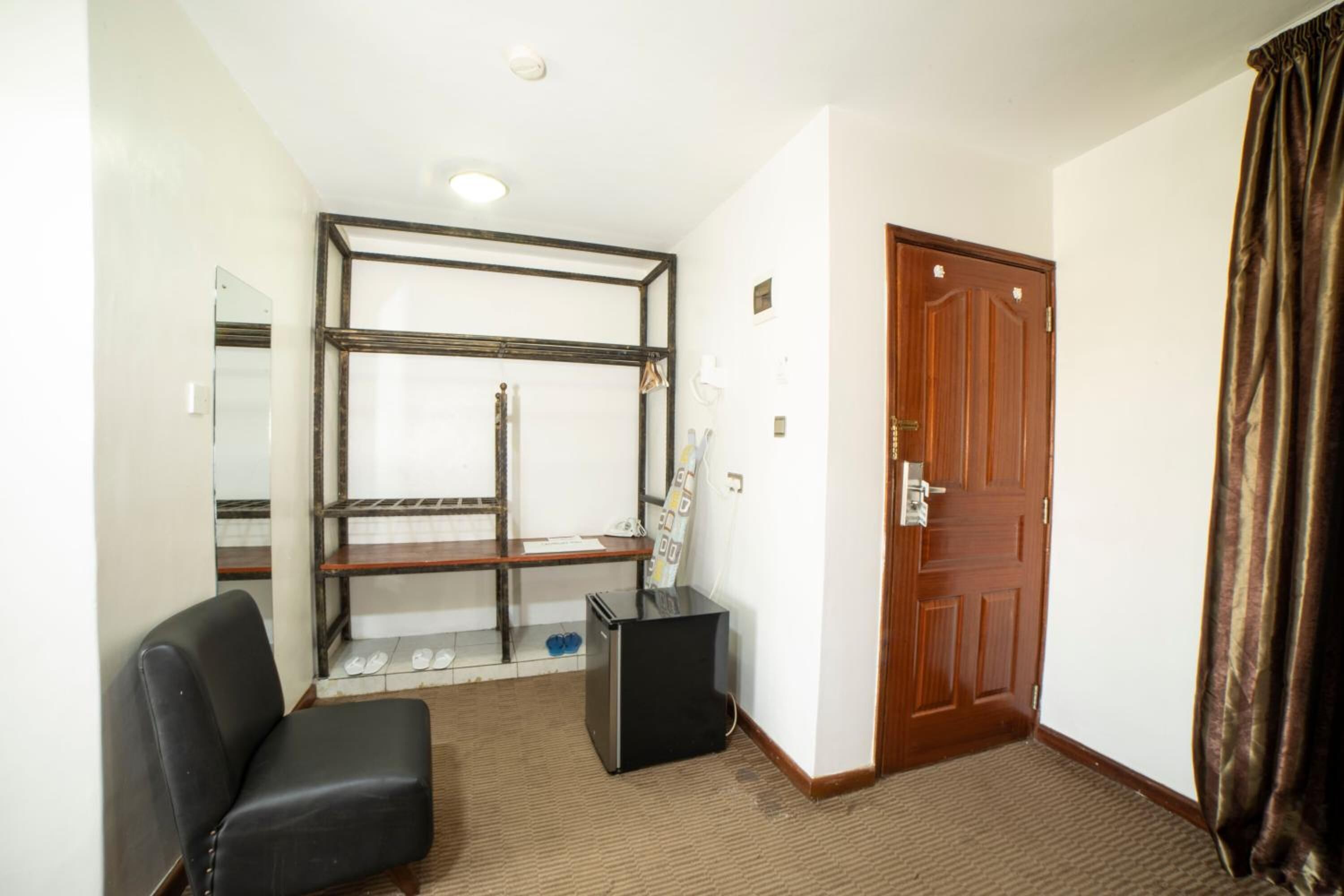 A hotel room with a wooden door, a black chair, a small black cabinet, and a mirror on the wall.
