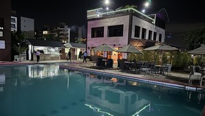 Area piscina di notte con un edificio sullo sfondo, posti a sedere all'aperto e un bar.