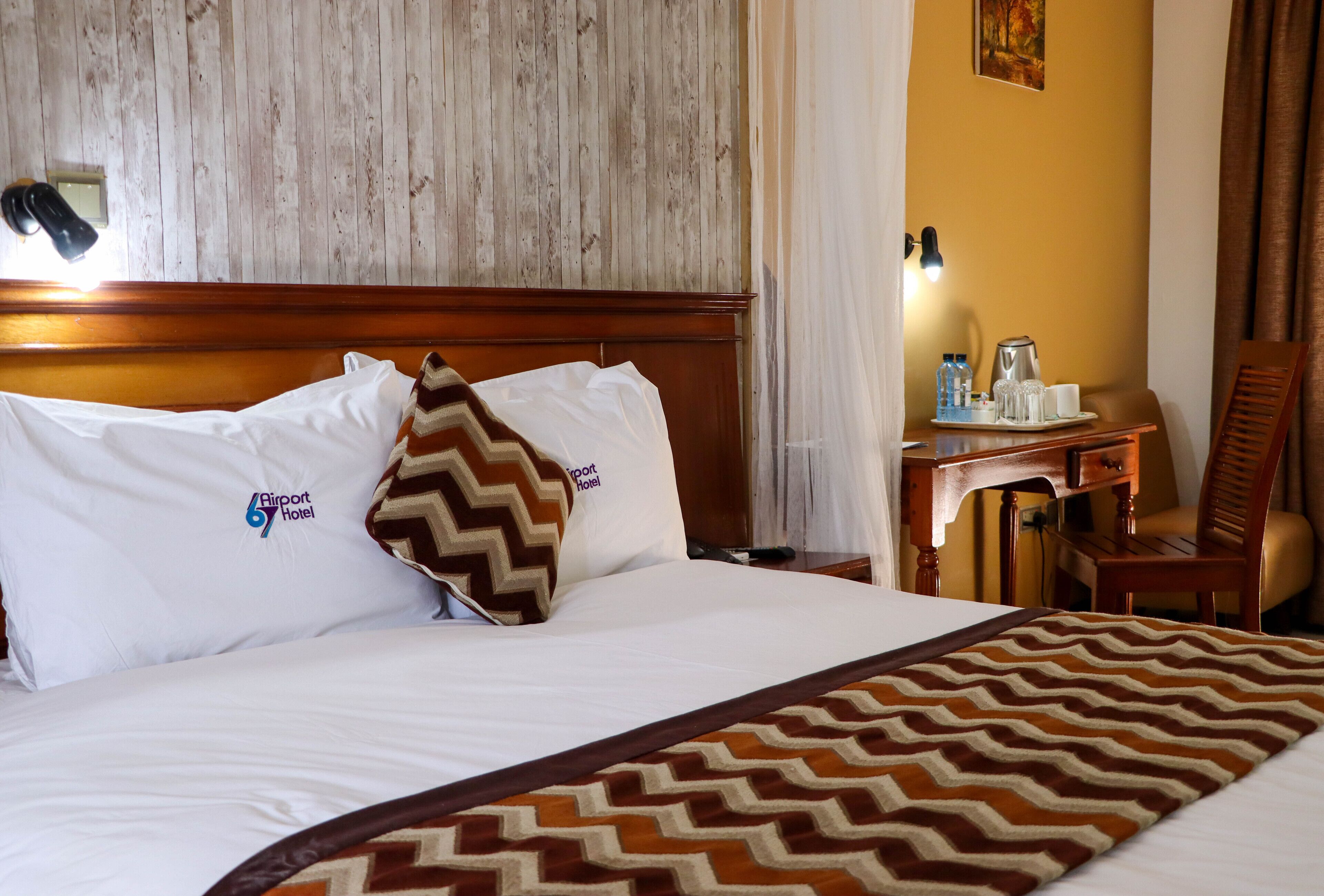 Ein Hotelzimmer mit einem hölzernen Kopfteil, einem Bett mit weißen Bettwäsche, einer gemusterten Tagesdecke, einem Schreibtisch mit Lampe und einem Stuhl.