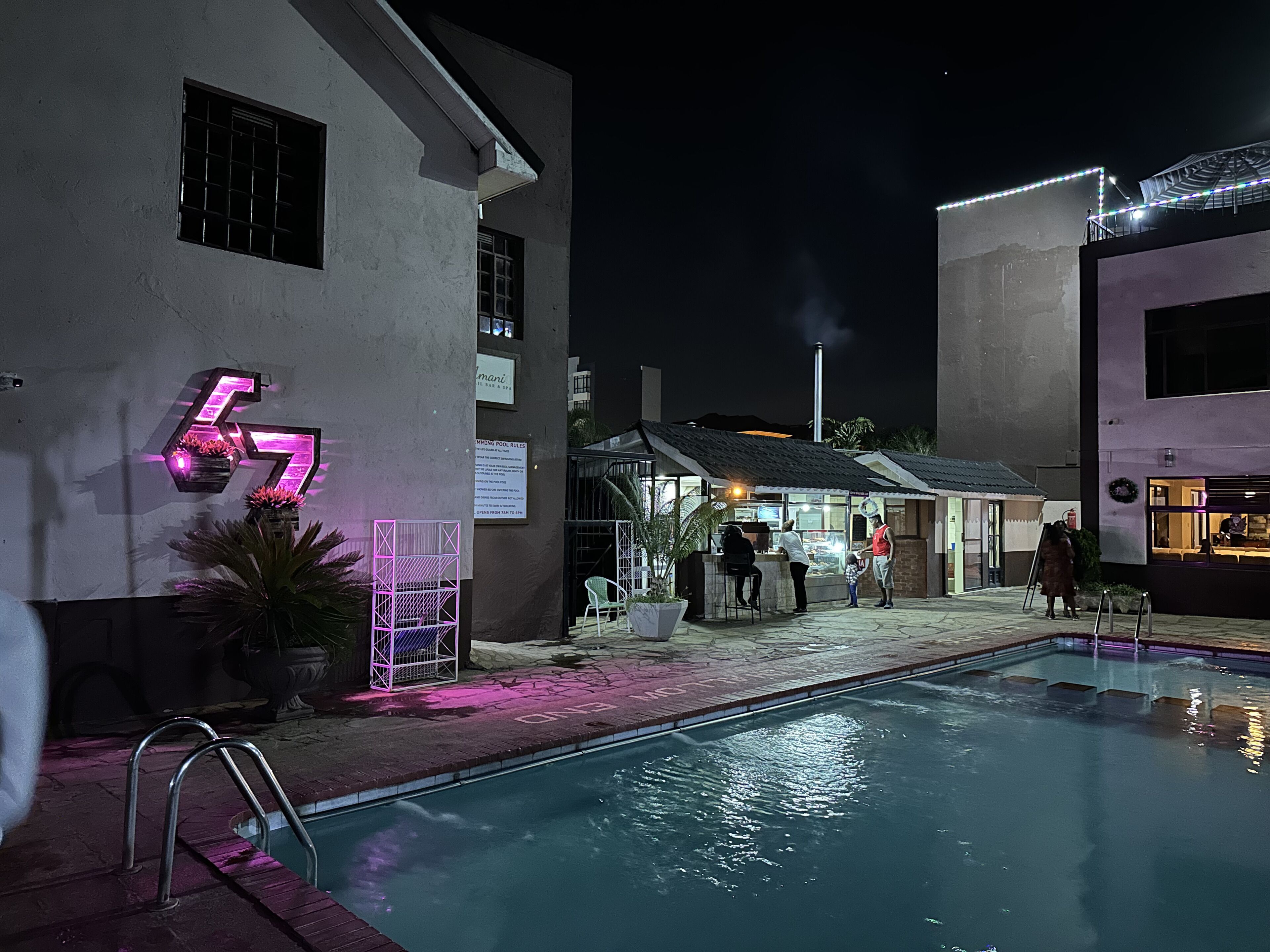 Ein Poolbereich mit einer Neonreklame, einer Poolbar und einem Gebäude mit einem Schild 'Schwimmbadregeln'.