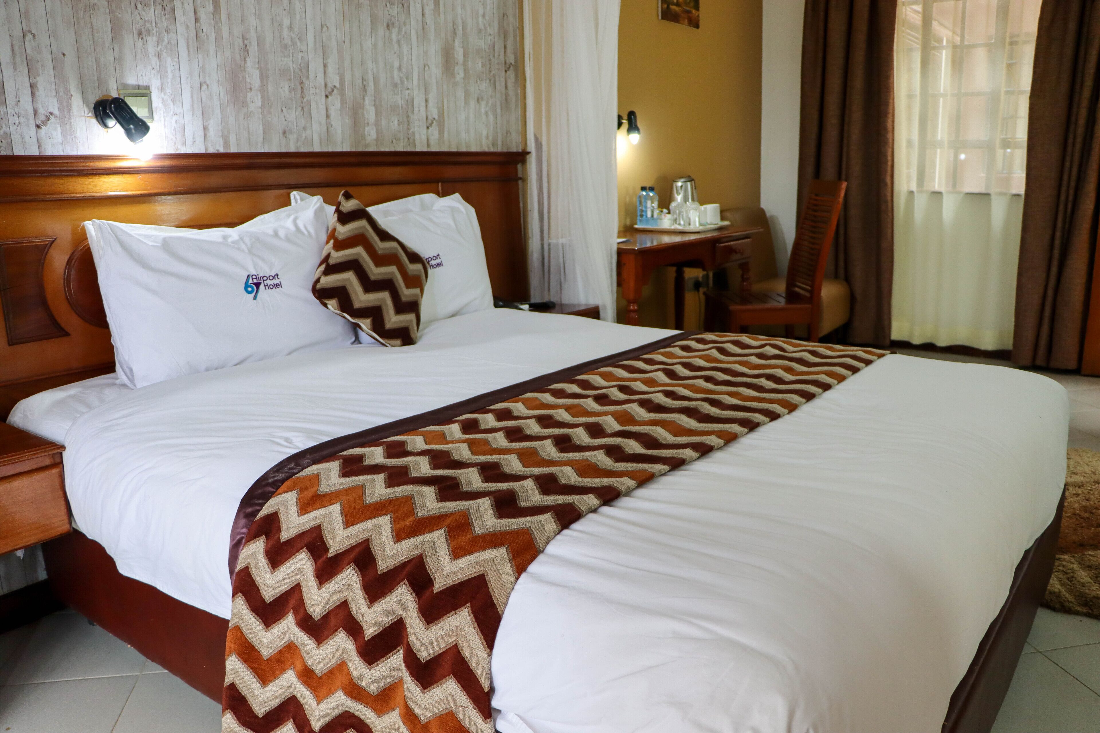 A hotel room with a large bed, a wooden headboard, a desk with a chair, a nightstand, and a window with curtains.