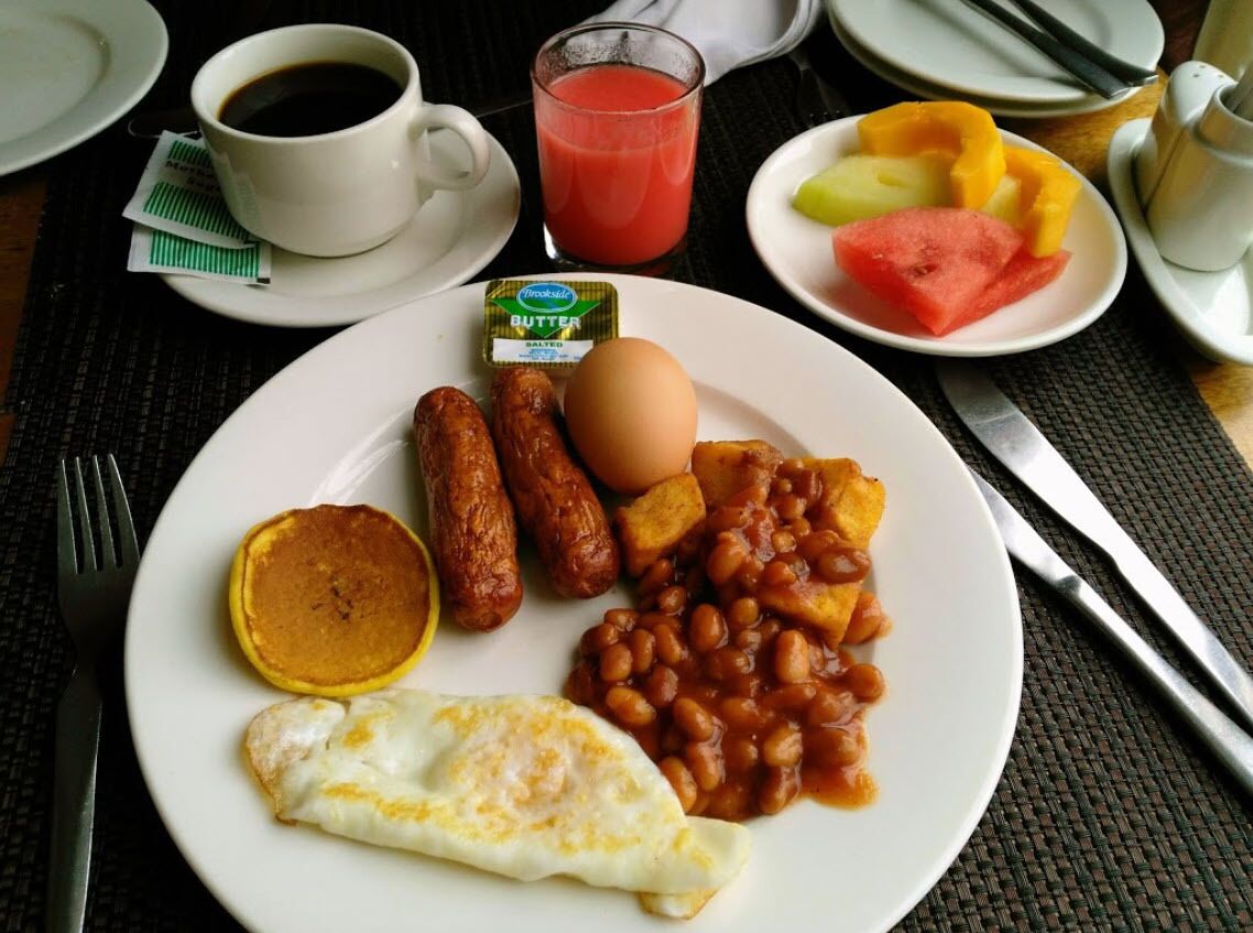 A breakfast plate with sausages, eggs, beans, and toast, accompanied by a cup of coffee and a glass of juice.