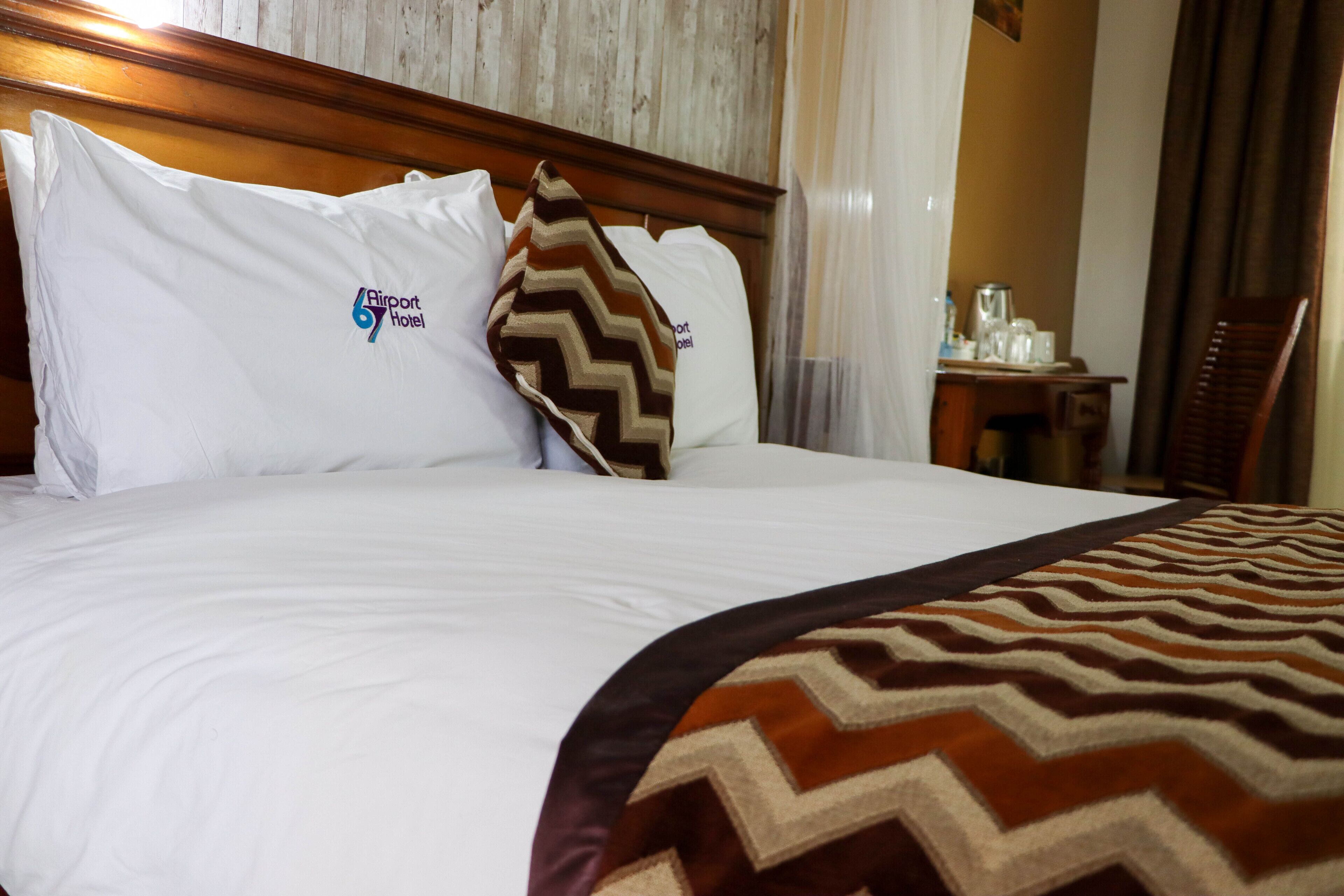 Un lit bien fait, avec du linge de lit blanc, un plaid à motifs chevrons marron et beige, et des oreillers ornés du logo de l’hôtel Airport 6.