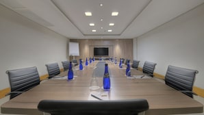 Un salón de conferencias con una larga mesa, sillas negras, botellas de agua azules y una pantalla de proyector.