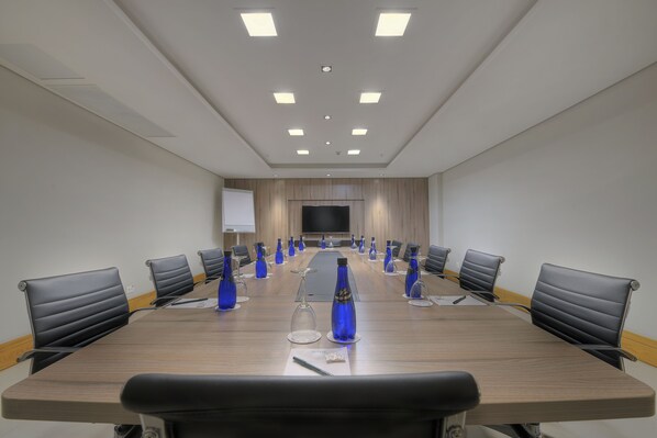 Une salle de conférence avec une longue table, des chaises noires, des bouteilles d’eau bleues et un écran de projection.
