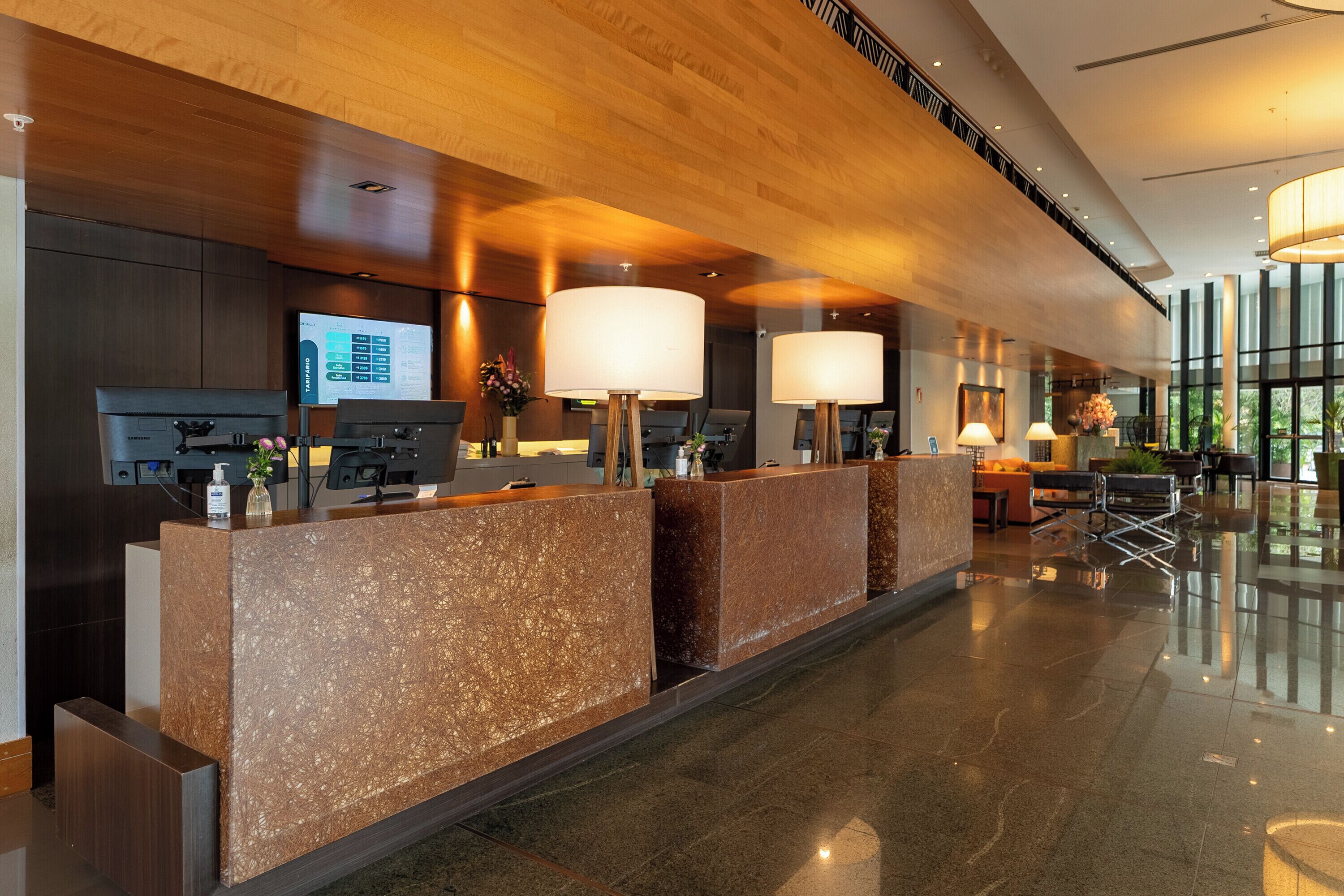 A hotel reception area with a modern design, featuring a reception desk, a digital screen, and a seating area with a view of the outdoors.
